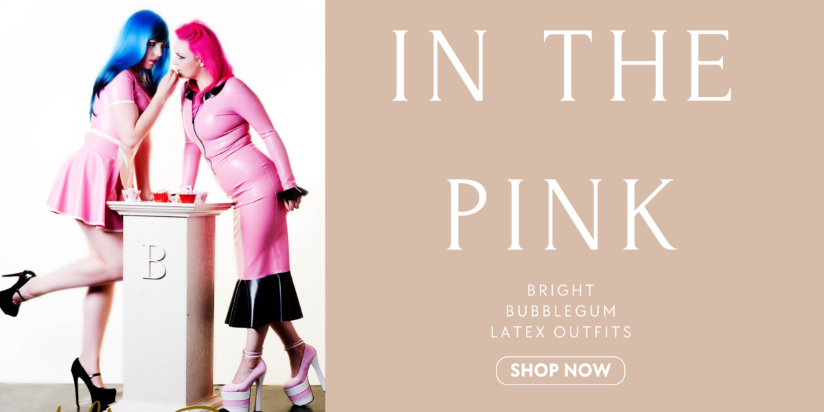 Two females are shown wearing bubble gum pink fetasia latex designs, One wears a two piece skirt suit. The jacket has a black zip, collar and cuffs. The other model wears a short sleeved latex swing dress. It has white trim detailing and a rounded edge apron.